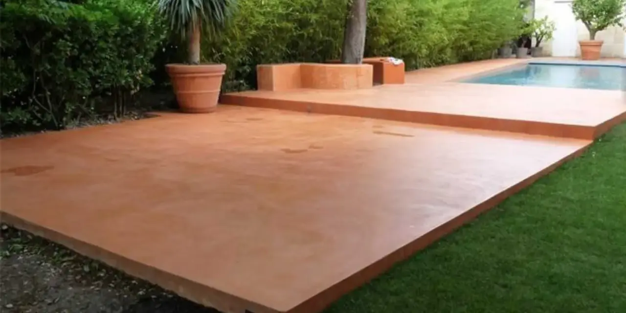 Une belle terrasse en béton coloré ocre, idéale pour un devis terrasse béton. Entourée de verdure et d'une piscine.
