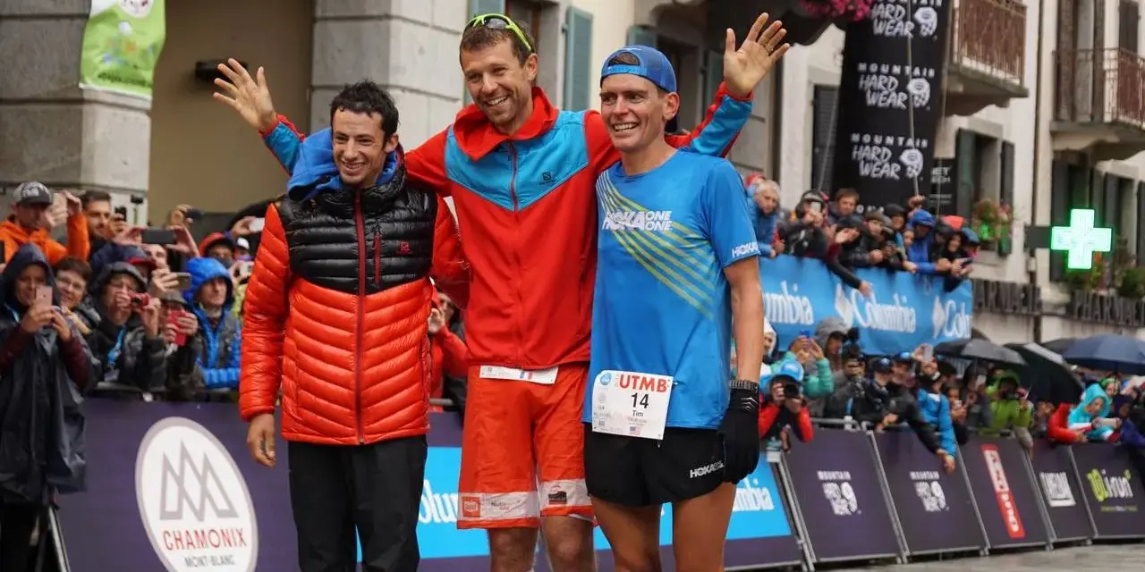 Trzech biegaczy na podium po ultra-trail du mont-blanc. Jeden w pomarańczowej kurtce, drugi w czerwonej, trzeci w niebieskiej koszulce.