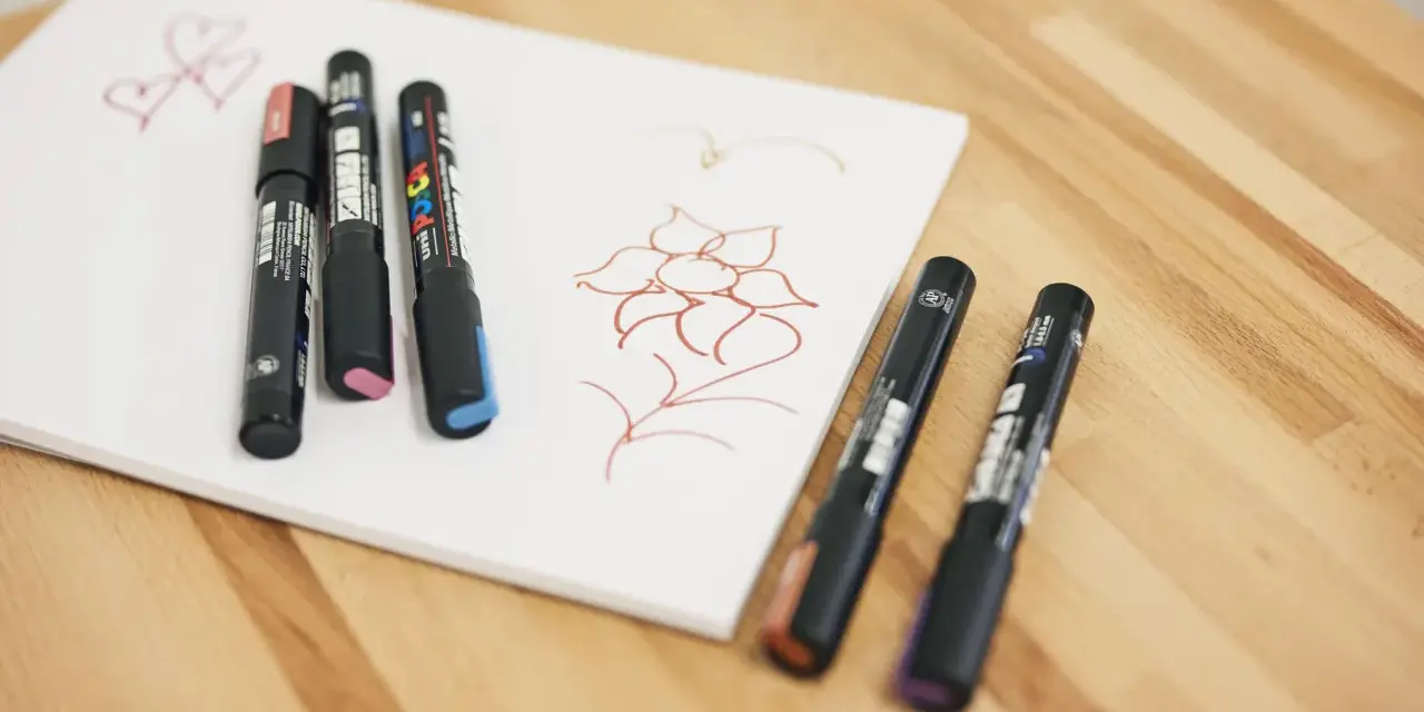 Några Posca-pennor ligger på ett vitt papper med en enkel blomma och hjärtan ritade i rött.