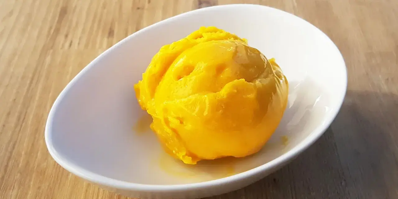 Ein leuchtend gelber Löffel Mango Sorbet in einer weißen Schale auf Holzhintergrund.