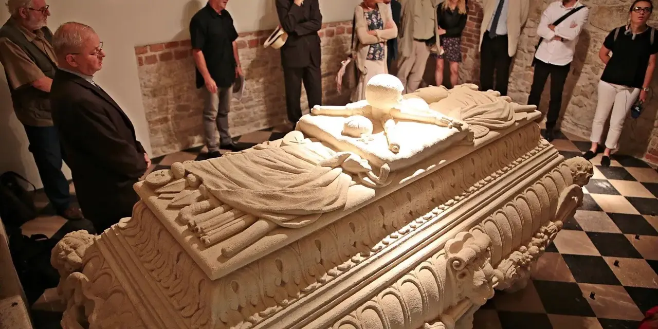 Grupa zwiedzających ogląda kamienny sarkofag w wawel krypty. Kamienne rzeźbienia zdobią jego bok.