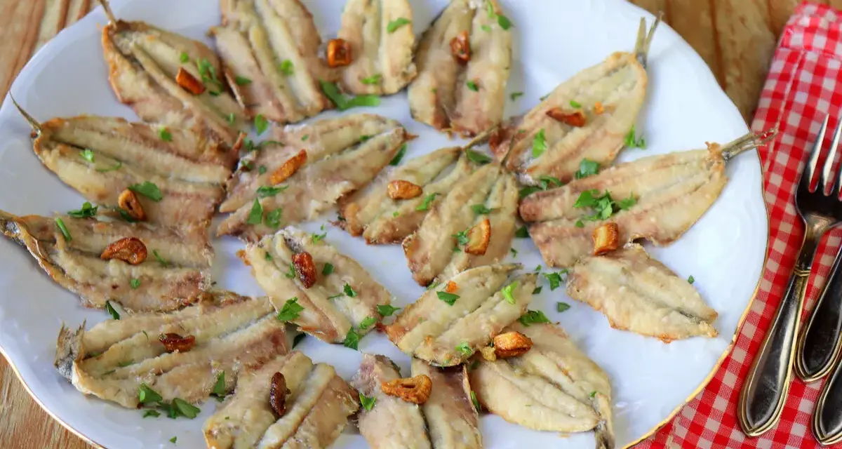 Un plato blanco con boquerones fritos, dorados y crujientes, espolvoreados con perejil fresco y trocitos de ajo.