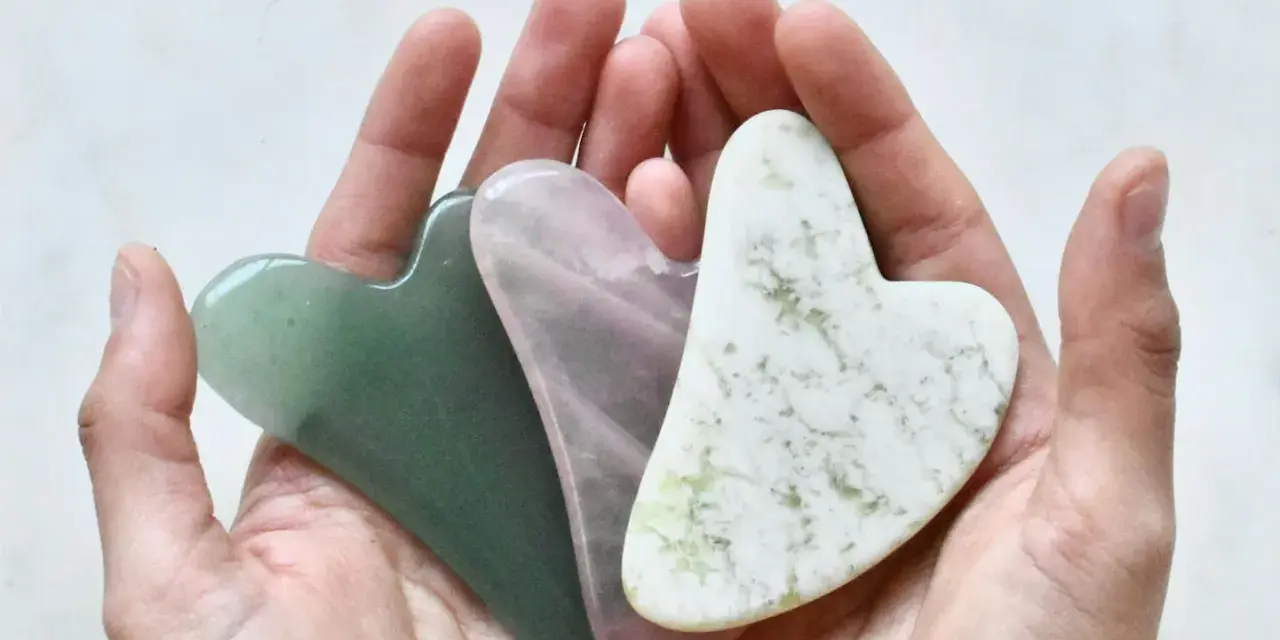 Drei herzförmige Gua Sha Steine in Grün, Rosa und Weiß liegen in einer Hand. Ideal für Gua Sha gegen Falten.