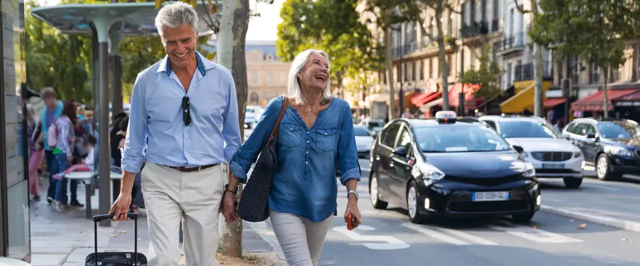 Uśmiechnięta para seniorów spaceruje po mieście z walizką, obok przejeżdżają samochody.