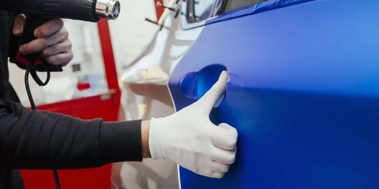 Ein Handwerker erwärmt mit einem Heißluftföhn eine blaue Folie, die auf ein Auto aufgebracht wird. Die auto folieren kosten sind hierbei ein wichtiger Faktor.