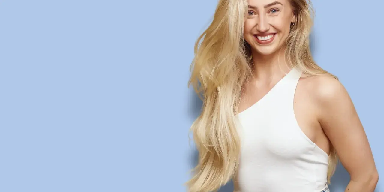 En leende kvinna med långt, blont hår som ser ut att bleka håret naturligt. Hon bär en vit topp och jeans mot en ljusblå bakgrund.