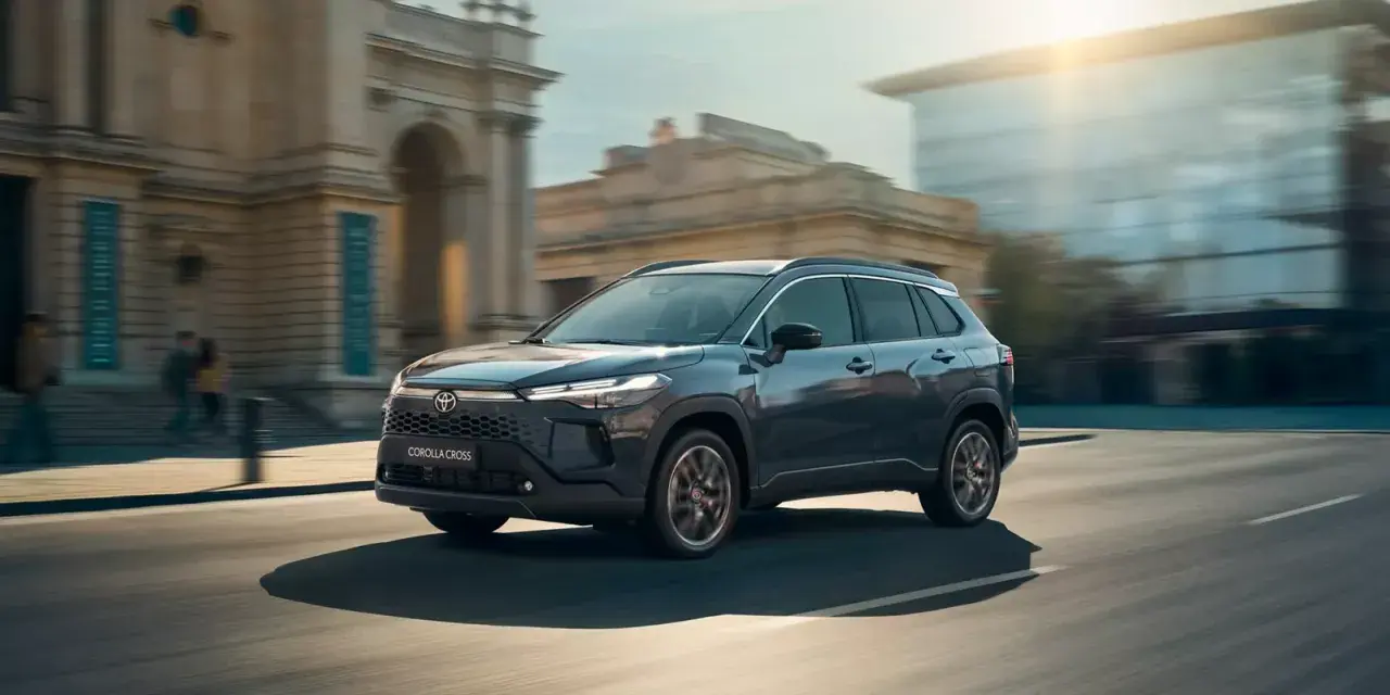 Ciemnoszary Toyota Corolla Cross Hybrid sunie ulicą miasta, odbijając światło słoneczne od nowoczesnych budynków.