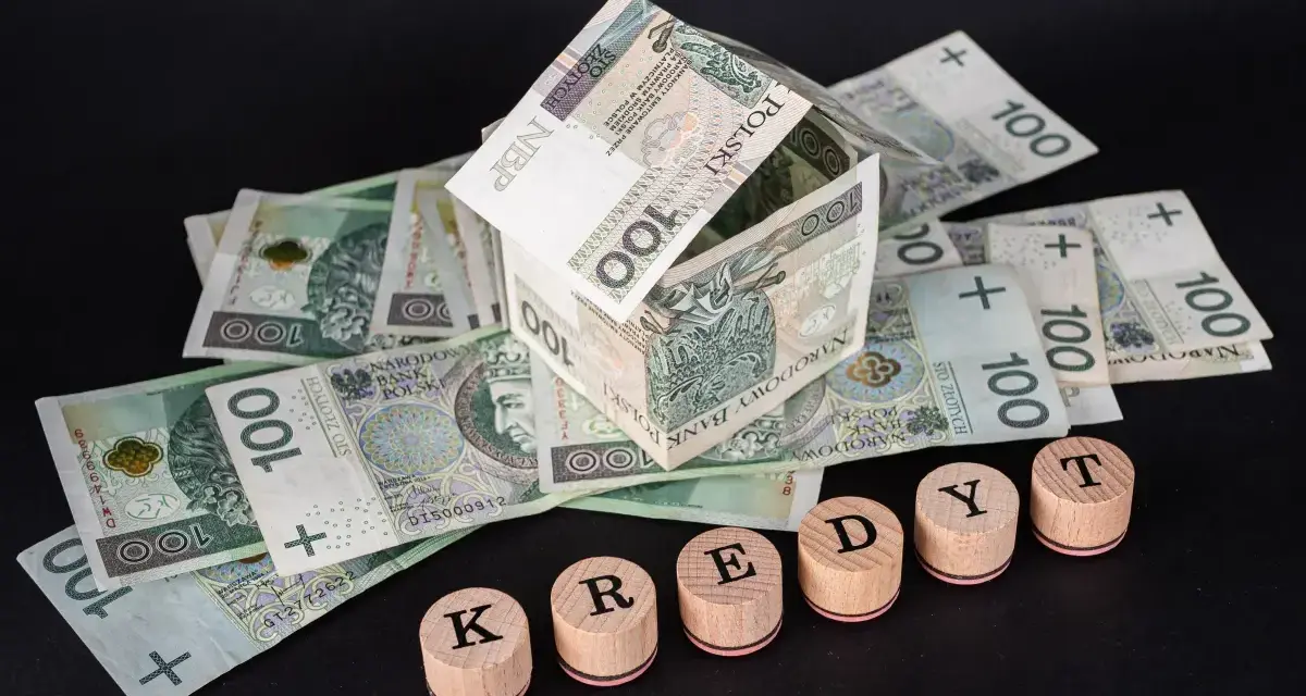 Dom z banknotów 100 zł symbolizuje marzenia o własnym lokum. Drewniane pieczątki z literami K, R, E, D, Y, T układają się w słowo "KREDYT", sugerując potrzebę finansowania lub **restrukturyzacja kredytu**.