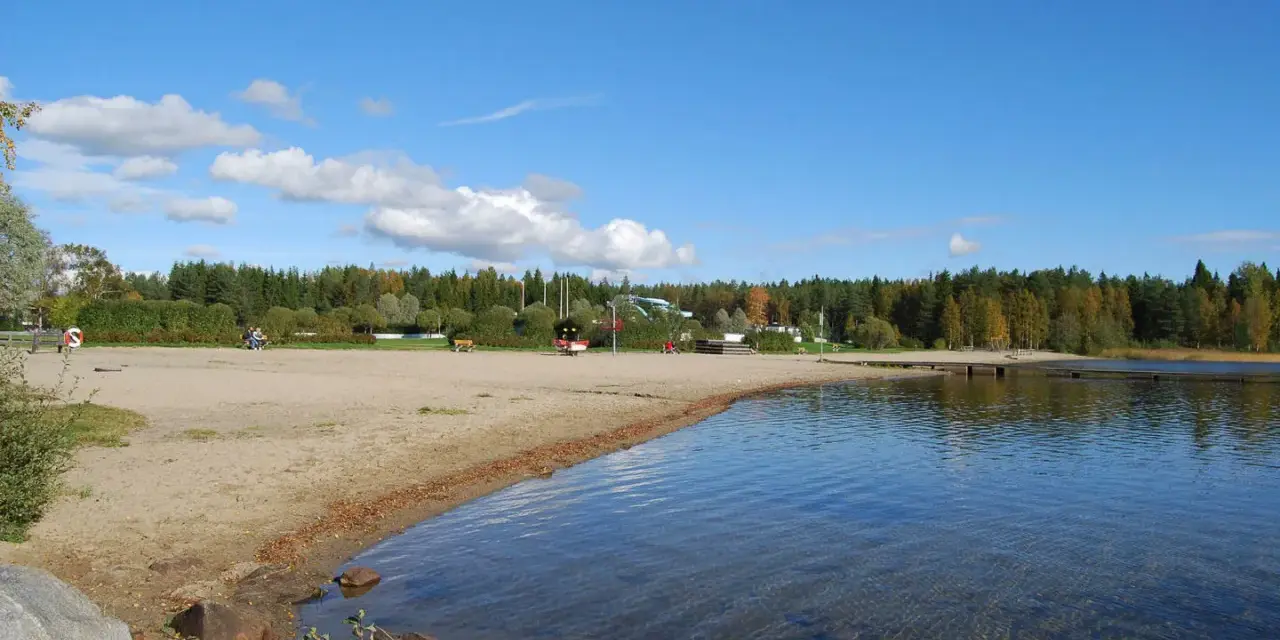 Solig strand vid Nydala camping Umeå med klart vatten och grönskande skog i bakgrunden.