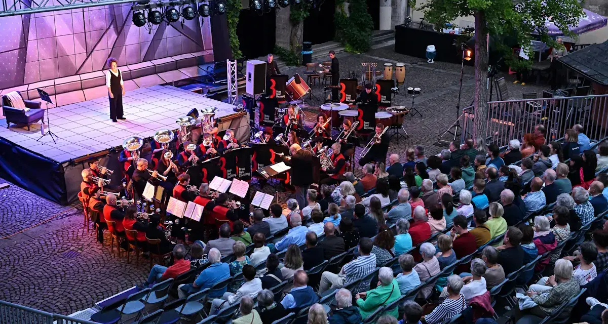 Eine Brass Band spielt auf einer Bühne, während ein Publikum auf Stühlen sitzt und zuhört.