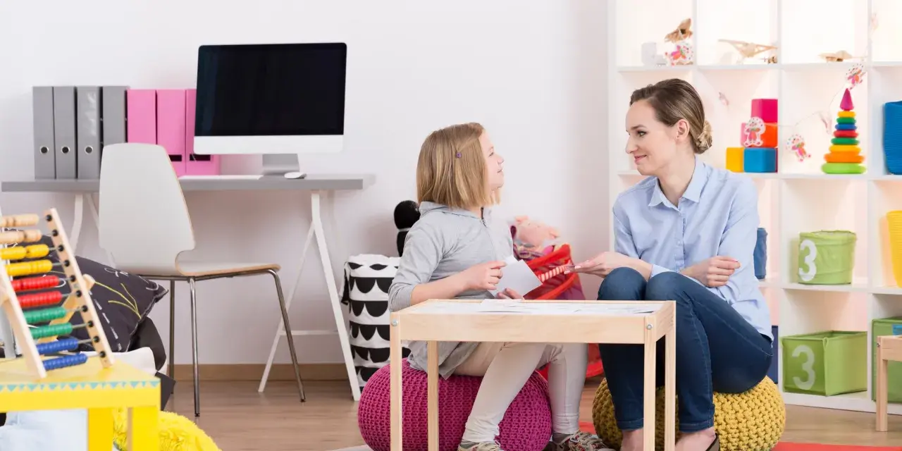 Terapeutka pracuje z dziewczynką, wspierając ją w terapii afazji u dzieci. W tle widać pomoce dydaktyczne i zabawki.