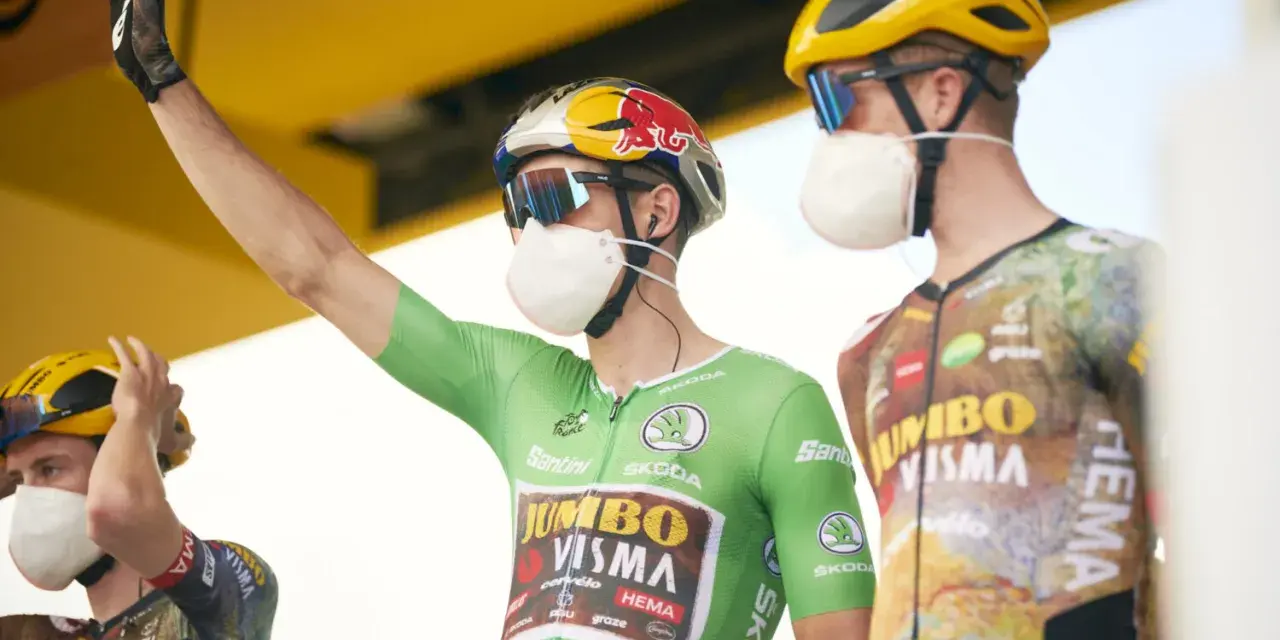 Cyclist in a green jersey, the symbol of the points classification leader at the Tour de France, raises his arm. He wears a mask and helmet.