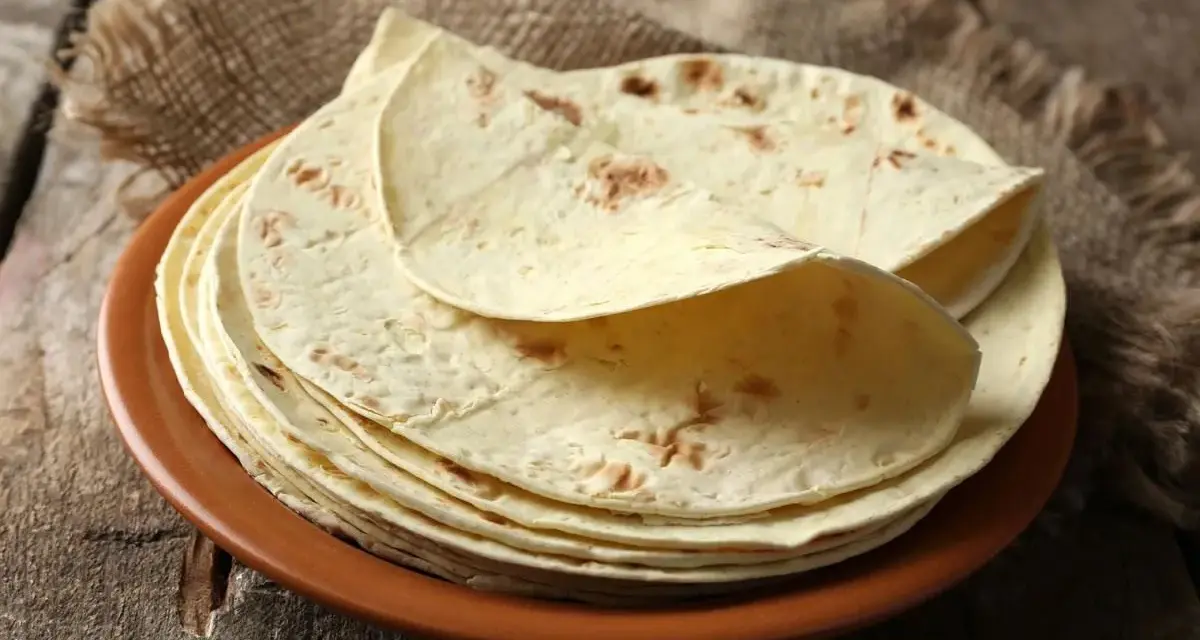 Stos cienkich, jasnych placków keto tortilla na brązowym talerzu, ułożonych na płótnie i drewnianym blacie.