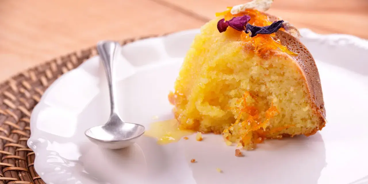 Un trozo de bizcocho de almendra de la abuela, decorado con ralladura de naranja y flores secas, servido en un plato blanco con una cucharilla.