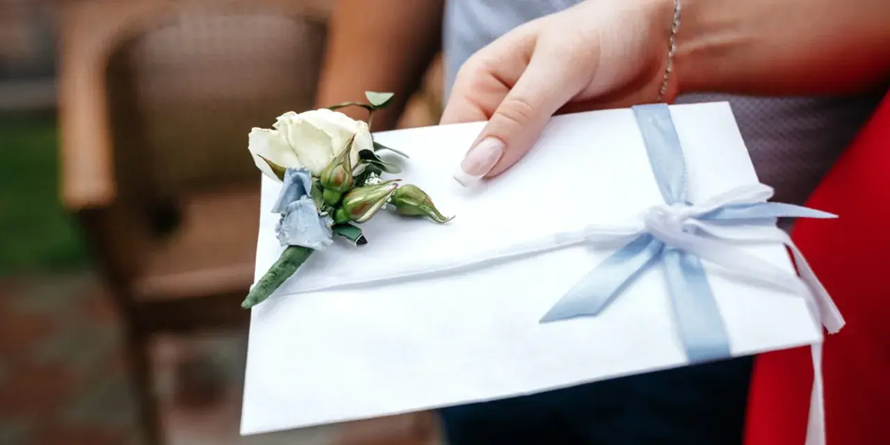 Dłoń trzyma białą kopertę z błękitną wstążką i ozdobnym kwiatkiem. Rozważasz skutki wręczenia pustej koperty lub zbyt małej kwoty? Opinie i etyka są tu kluczowe.