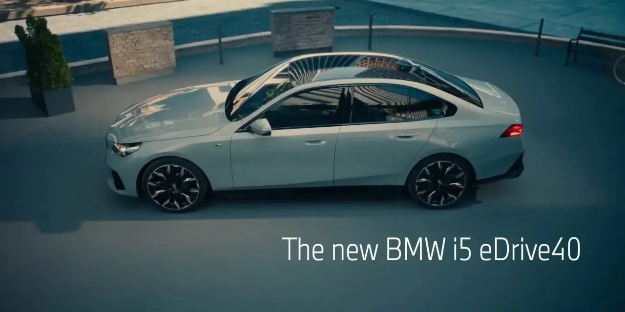 Jasnoszary sedan BMW i5 eDrive40 stoi na zewnątrz, z widokiem na basen i ławkę.