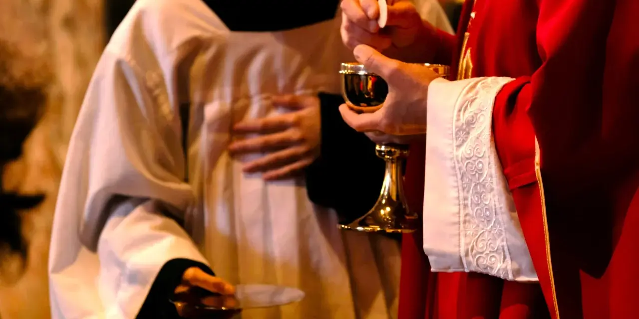 A priest in red vestments holds a chalice and host, while another in white vestments holds a paten. A moment of communion, perhaps a prayer to St. Benedict for healing.