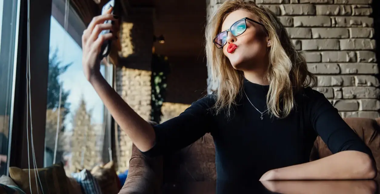 Kobieta w okularach robi sobie selfie, wyciągając usta do pocałunku. W jej spojrzeniu widać pewność siebie, graniczącą z narcyzmem.