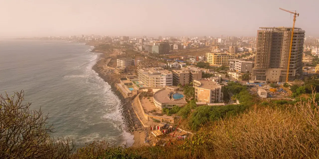 Widok na wybrzeże i rozwijające się miasto w **Afryce, Senegal**. Widać ocean, plażę, nowoczesne budynki i plac budowy z dźwigiem.