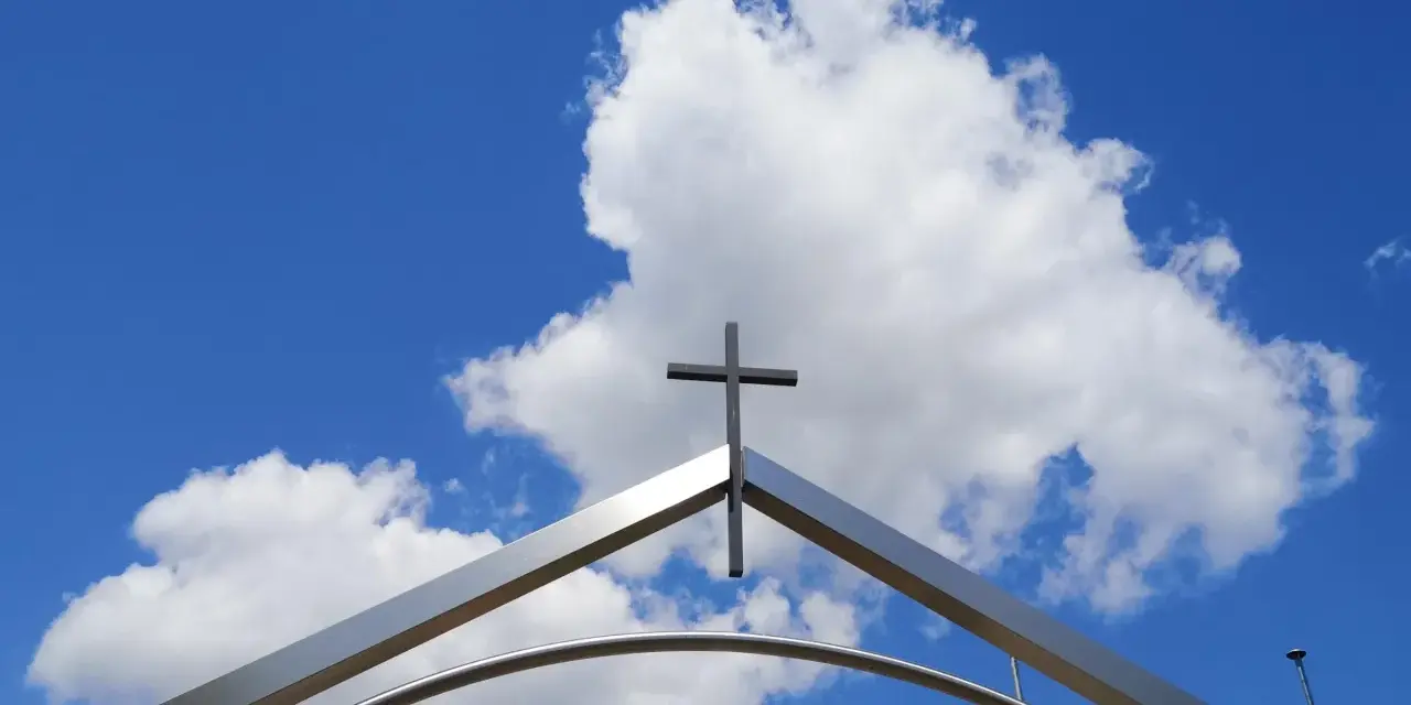 Ein Kreuz ragt über einem modernen Torbogen in den blauen Himmel mit Wolken. Ein starkes Christi Himmelfahrt Symbol.
