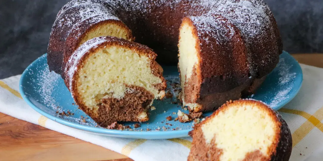 Saftiger weltbester Marmorkuchen, mit Puderzucker bestäubt, auf blauem Teller. Ein Stück ist abgetrennt und zeigt die köstliche Marmorierung.