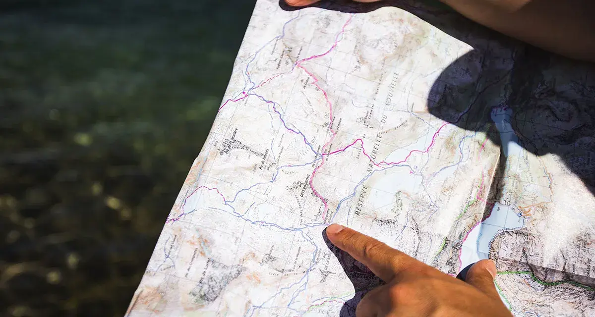 Une main pointe un itinéraire sur une carte topo détaillée, près d'une étendue d'eau.