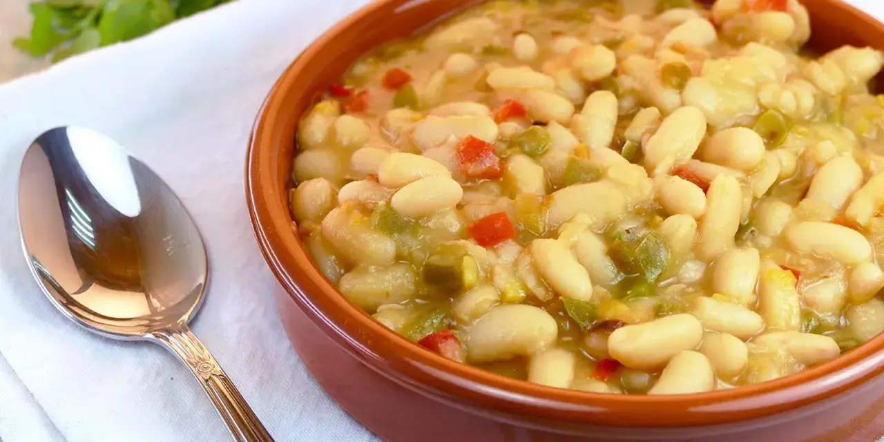 Un cuenco de barro lleno de alubias blancas con verduras, como pimientos rojos y verdes. Una cuchara plateada descansa a la izquierda.