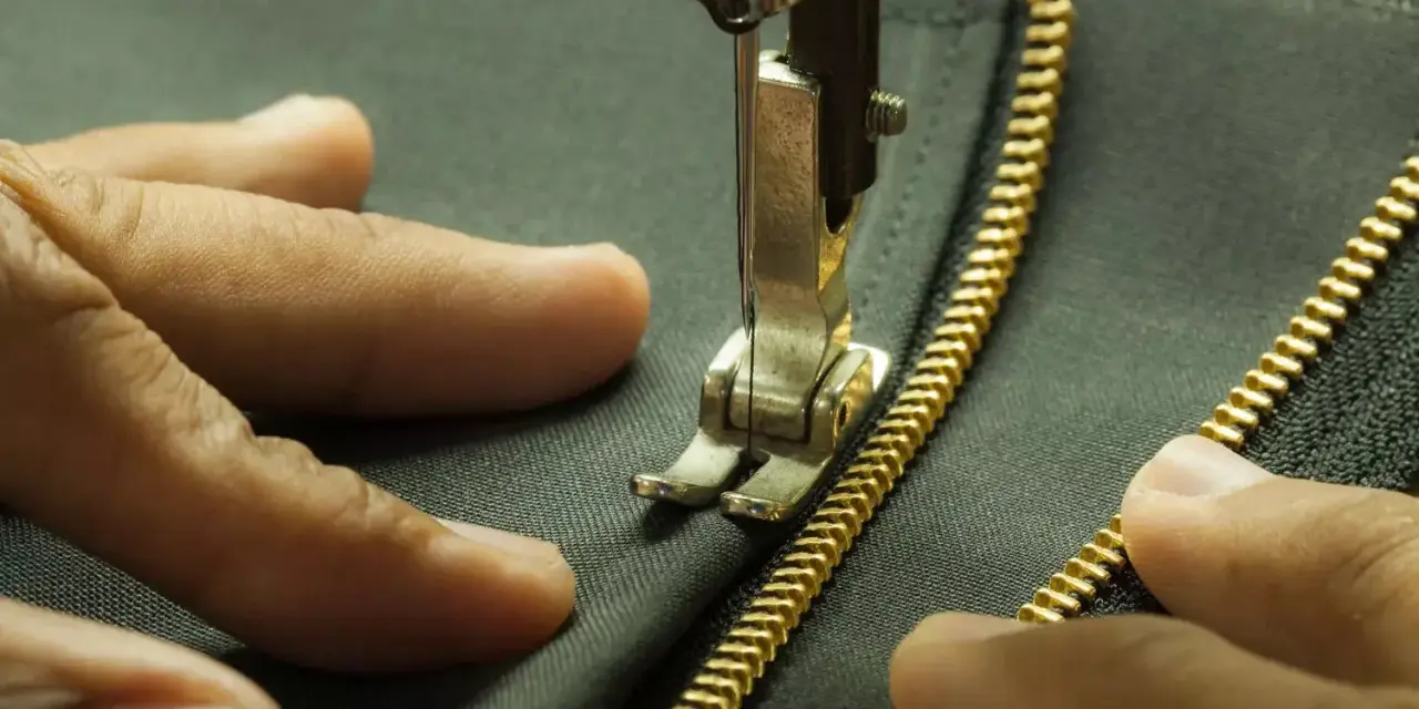 Main tenant une fermeture éclair dorée, une aiguille de machine à coudre s'apprête à coudre une fermeture éclair à la main sur un tissu sombre.