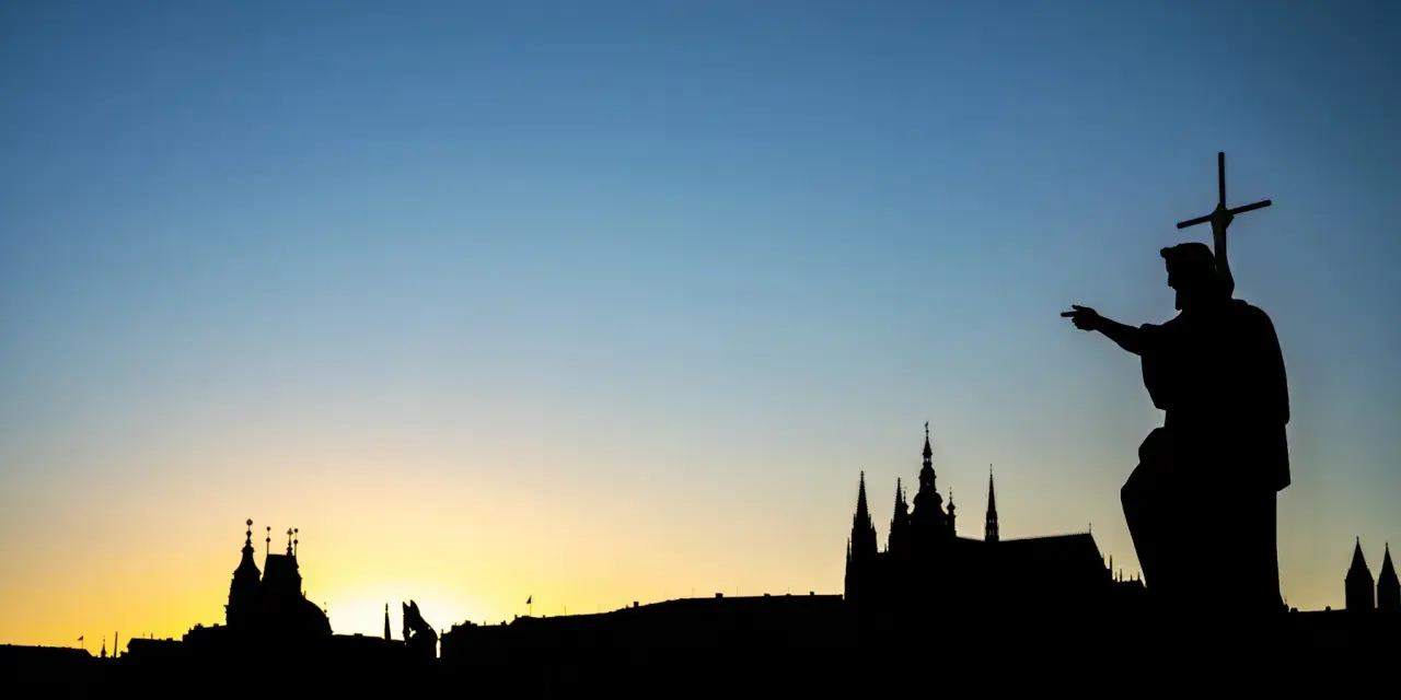 Silhouette d'une statue de saint pointant vers un paysage urbain au coucher du soleil. Le ciel bleu dégradé est le centre d'intérêt de l'image.