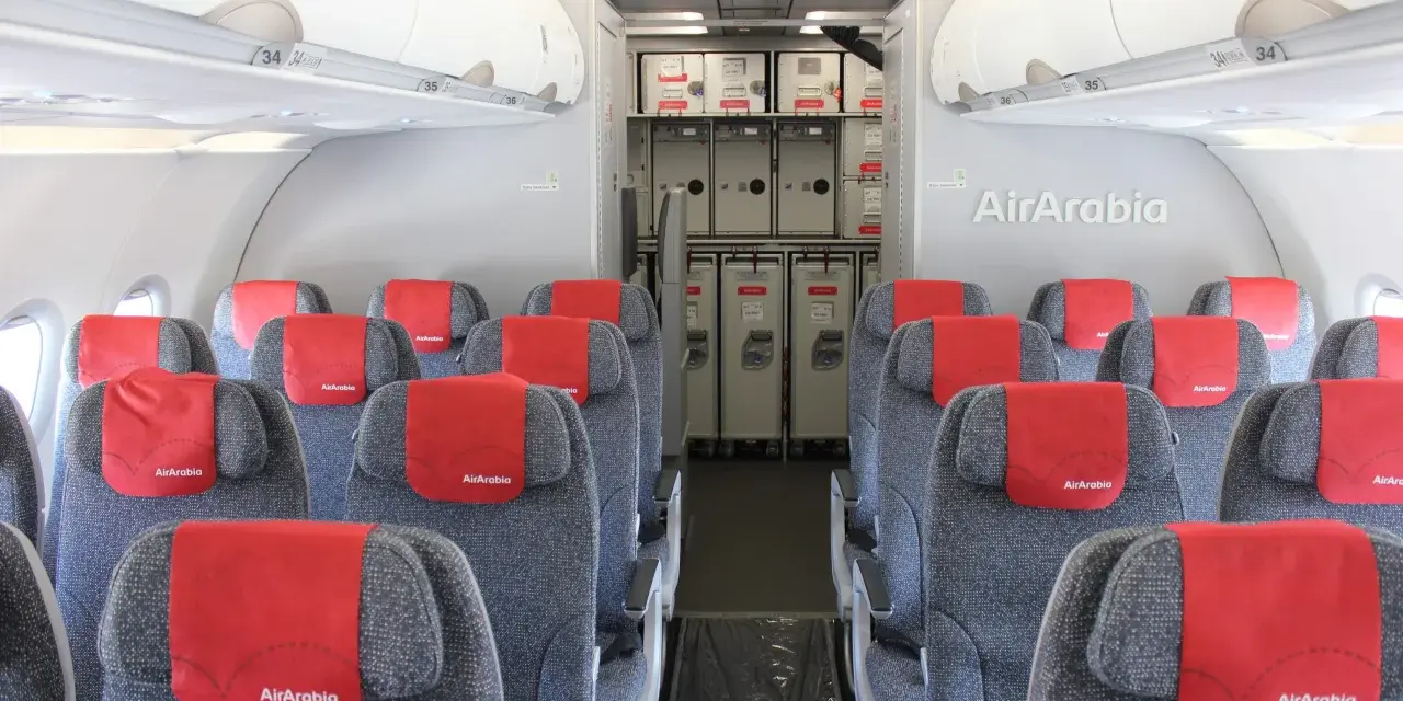 Wnętrze samolotu Air Arabia z rzędami szarych siedzeń i czerwonymi zagłówkami. W tle widać wózki pokładowe. Dobre **air arabia opinie** potwierdzają komfort podróży.
