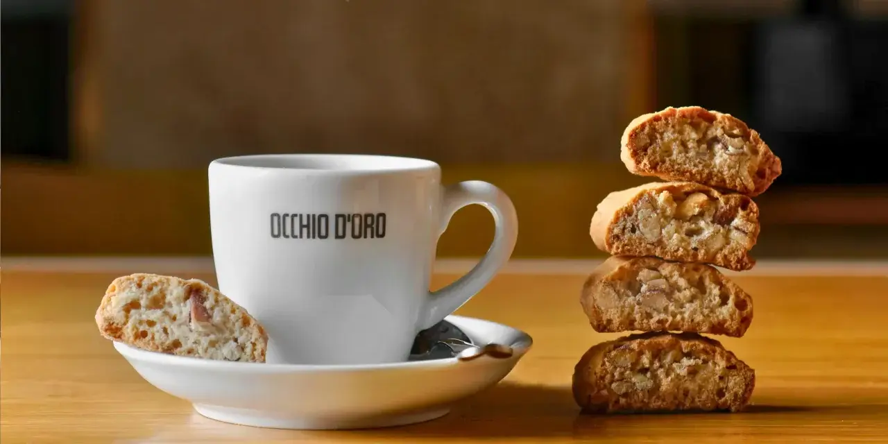 Ein weißer Kaffeebecher mit "OCCHIO D'ORO" Schriftzug steht neben einem Stapel Cantuccini. Ein Cantuccino liegt auf der Untertasse. Dieses Bild inspiriert zum besten Cantuccini Rezept.