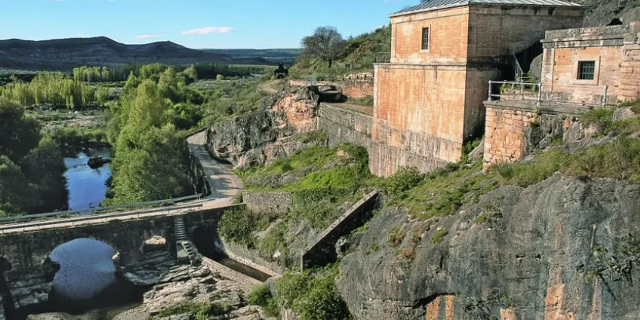 El portón de la oliva, un antiguo edificio de piedra junto a un río y un puente de arco, se alza sobre un paisaje rocoso y verde.