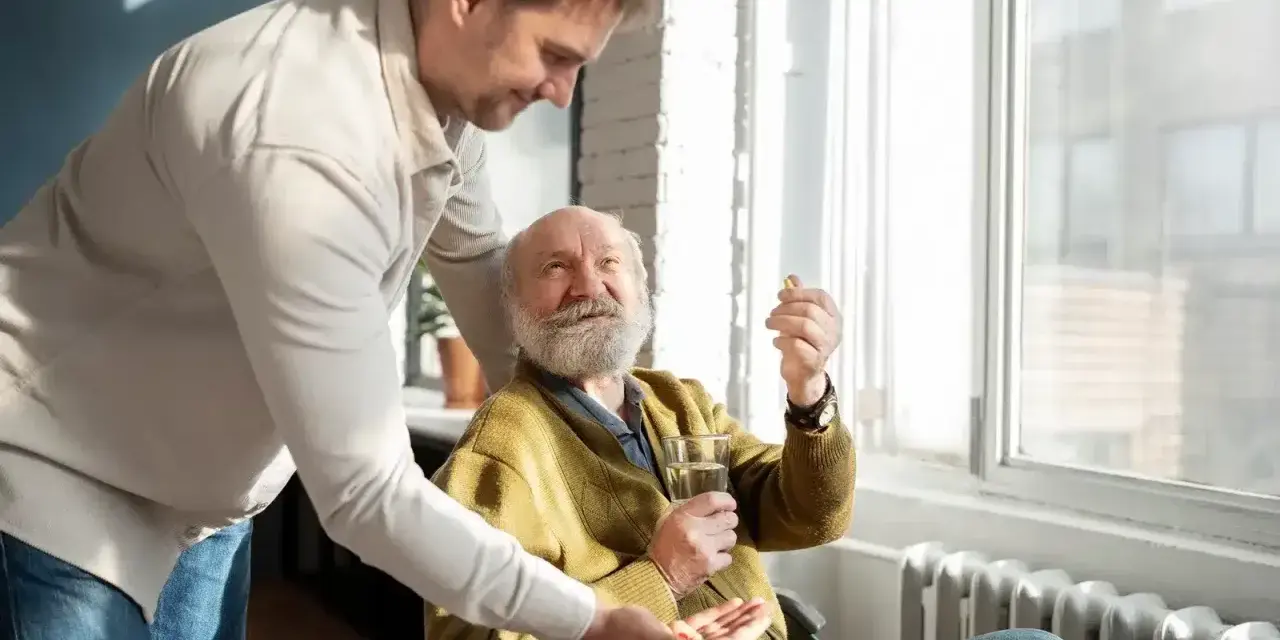 Mężczyzna w średnim wieku podaje starszemu mężczyźnie w wózku inwalidzkim tabletkę i szklankę wody.