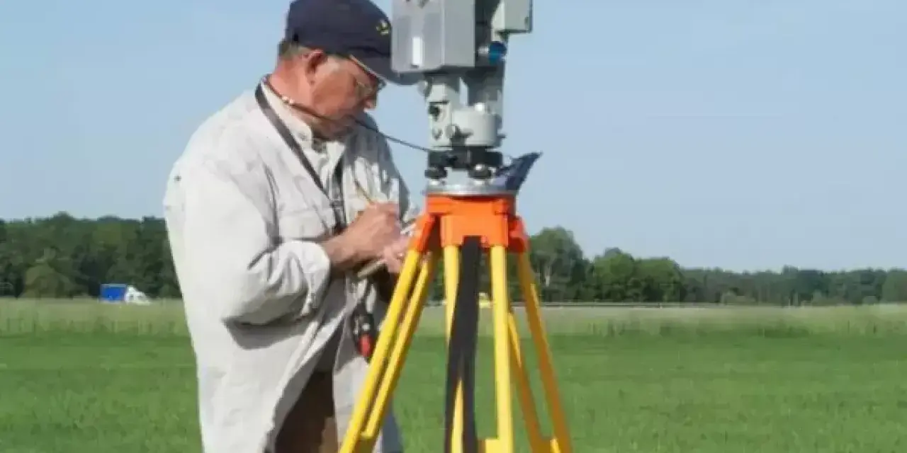 Geodeta precyzyjnie wyznacza granice działki za pomocą tachimetru na statywie, pracując na zielonym polu.