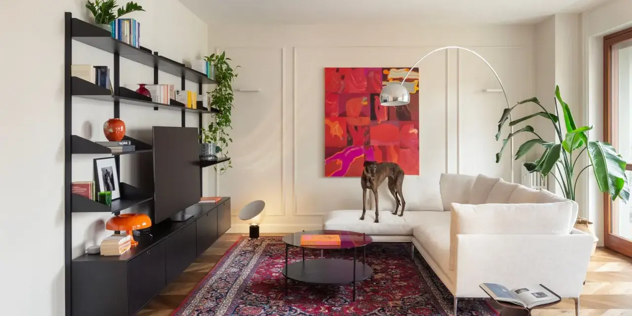 Un galgo se posa sobre un sofá blanco, rodeado de arte abstracto y un tapete persa. El **ruido visual** de los objetos decorativos y la pintura vibrante crea un ambiente ecléctico.