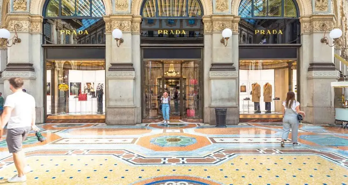 Ingresso del negozio **Prada** con vetrine eleganti e un mosaico colorato sul pavimento. Persone passeggiano davanti alla boutique.