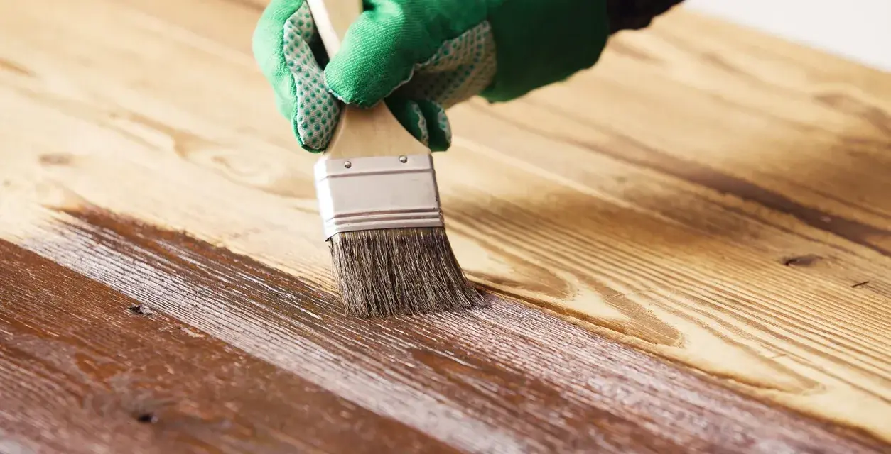 Mano guantata applica vernice protettiva con pennello per impermeabilizzare il legno, creando una finitura lucida.