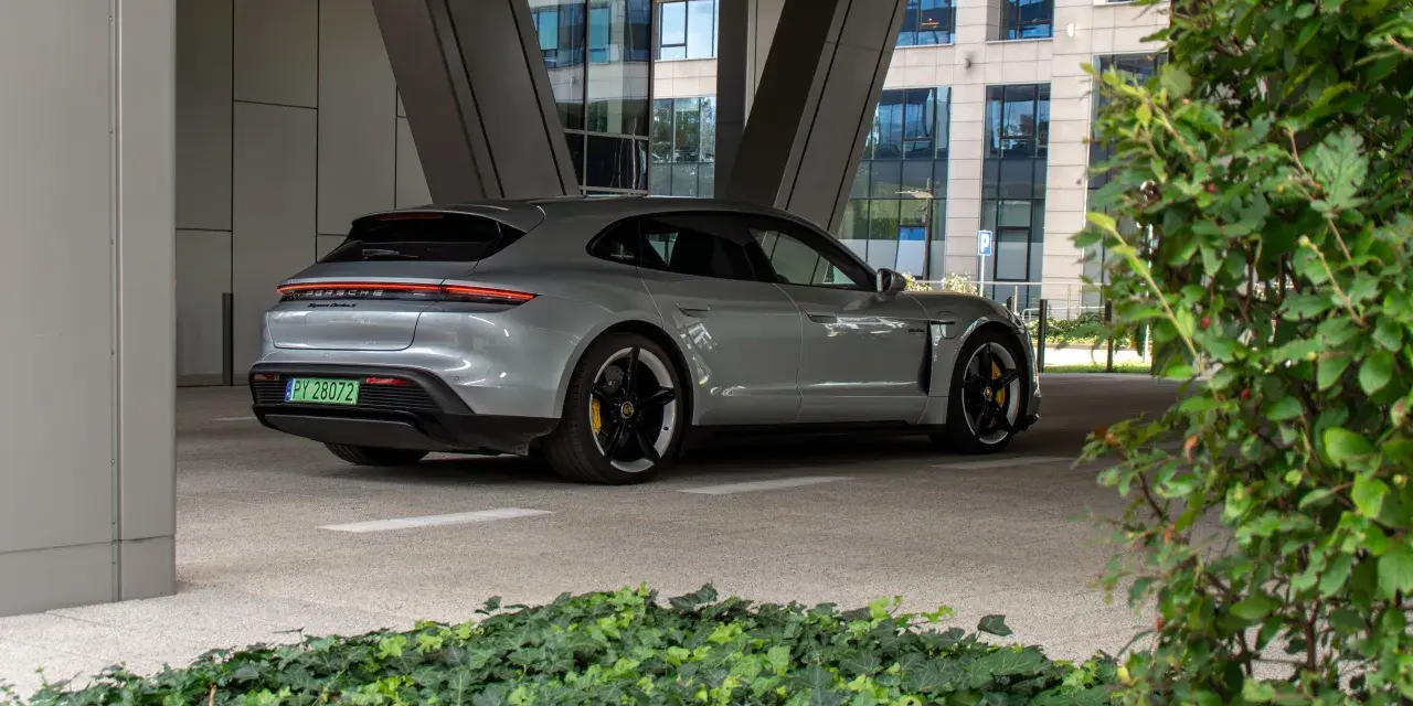 Szare Porsche Taycan Turbo S stoi pod zadaszeniem, z widokiem na nowoczesny budynek i zieleń.