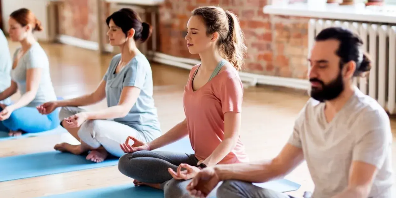 Menschen in einem Yogastudio praktizieren Stress-Meditation, sitzen im Lotussitz auf Matten und schließen die Augen.