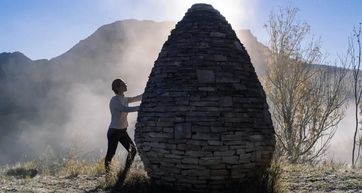 Une femme interagit avec une structure ovoïde en pierres, une œuvre de land art, dans un paysage brumeux et montagneux au lever du soleil.