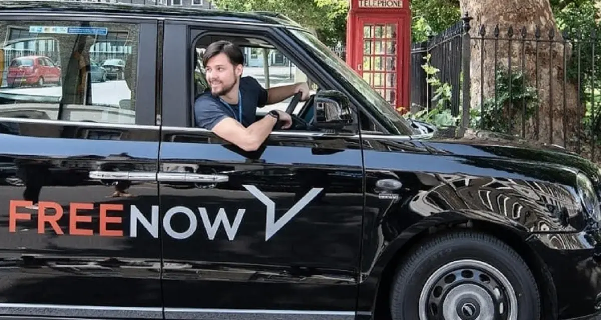Ein Mann fährt ein schwarzes freenow taxi. Im Hintergrund steht eine rote Telefonzelle.