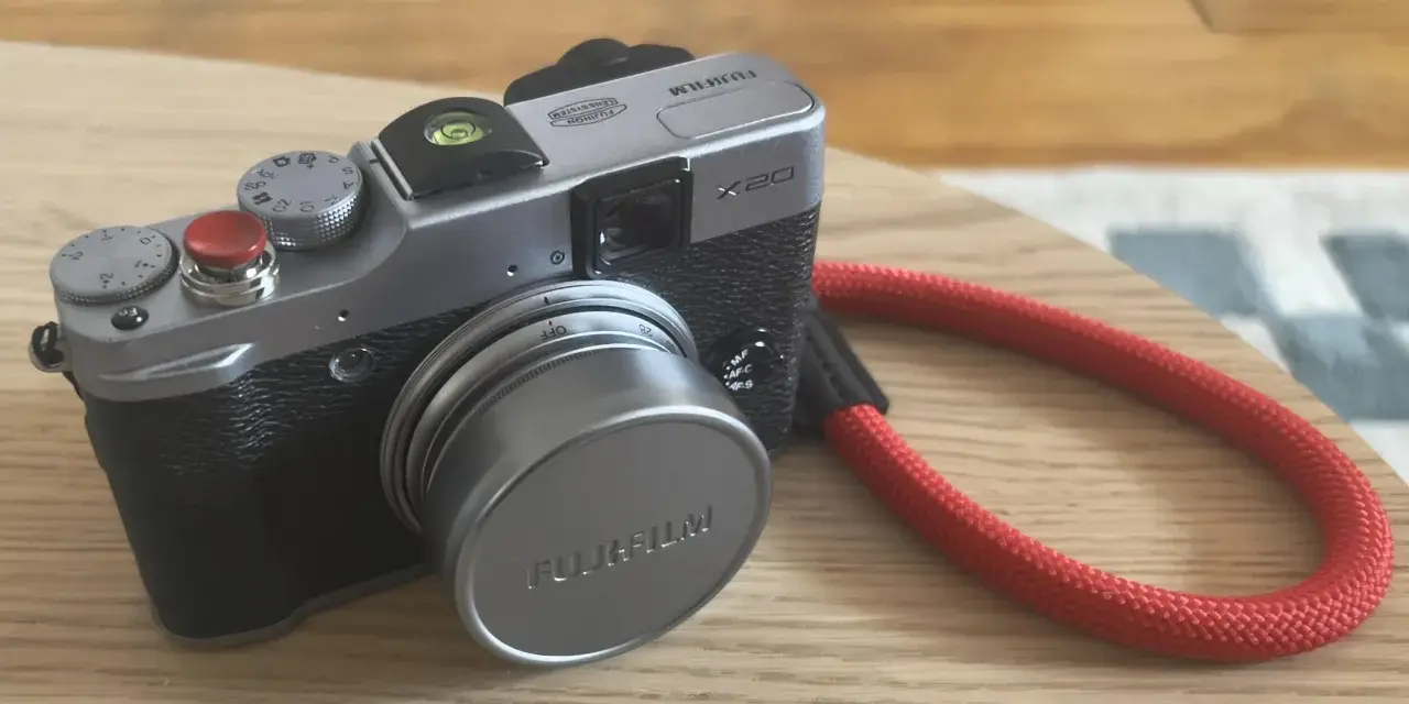 Srebrny aparat Fujifilm X20 z czerwoną smyczą leży na drewnianym blacie.