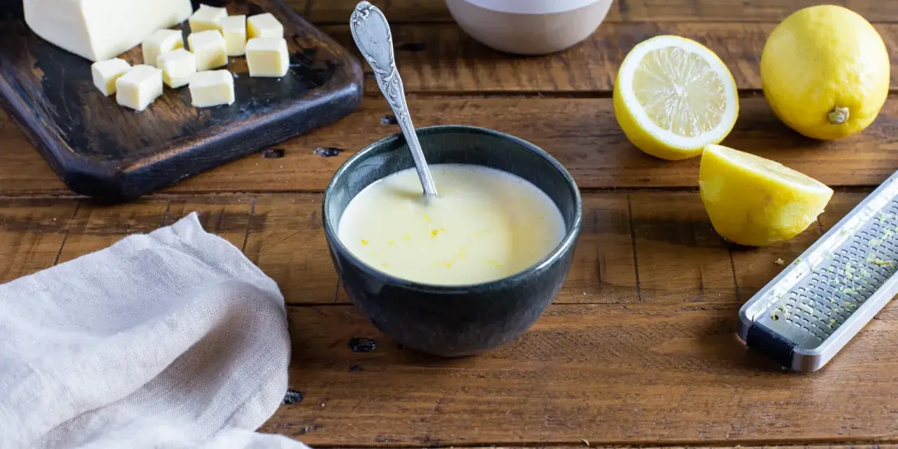 Un bol de sauce beurre citron, avec des citrons, du beurre coupé en dés et une râpe à zester sur une table en bois.