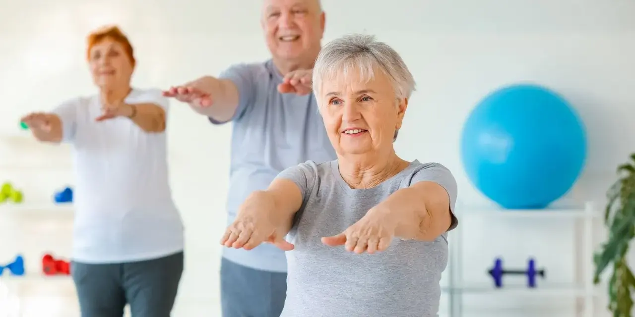 Trois seniors souriants pratiquent la gym douce, bras tendus, dans une salle lumineuse avec un ballon bleu en arrière-plan.
