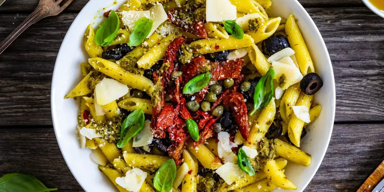 Penne con pesto, pomodori secchi, olive nere, capperi e scaglie di parmigiano. Un piatto colorato e saporito.
