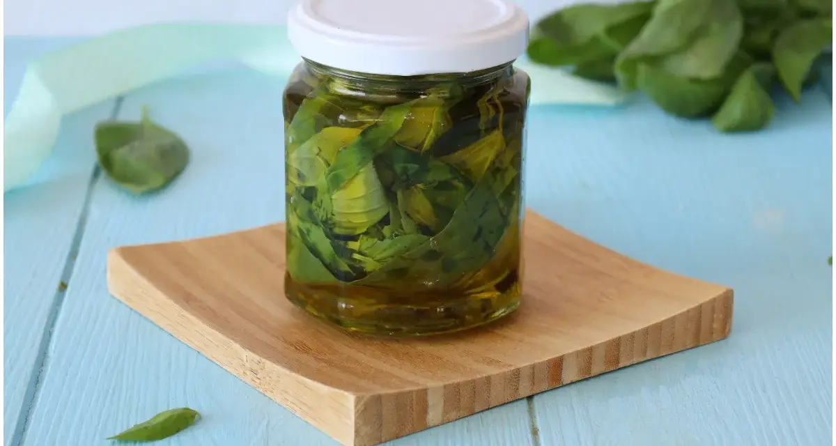 Un barattolo di olio al basilico fatto in casa, con foglie fresche immerse nell'olio dorato, su un tagliere di legno.