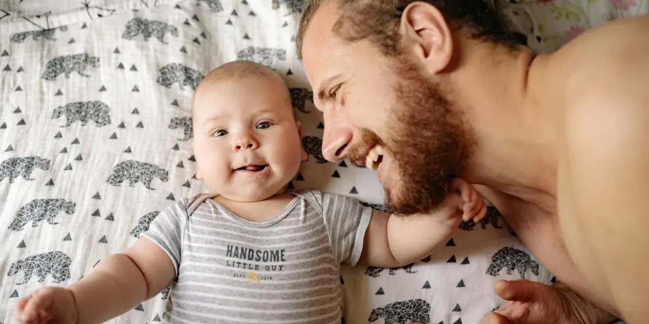 Ein lächelnder Vater und sein Baby liegen auf einer Decke mit Bärenmuster. Die Frage, ob Autismus vererbbar ist, beschäftigt viele, und die Bindung zwischen Vater und Kind ist ein wichtiger Aspekt.