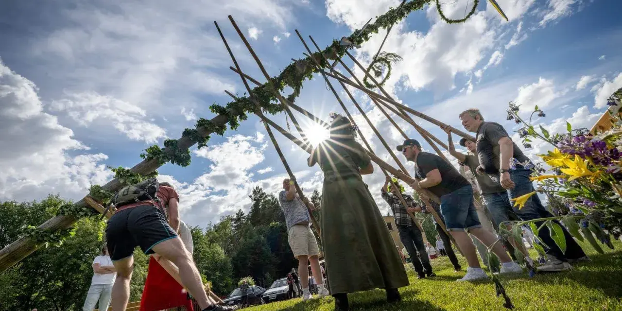 Midsommarstången reses av en grupp människor, en traditionell del av hur firar man midsommar. Solen skiner genom molnen.