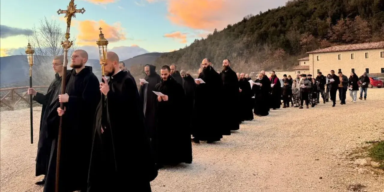Monks in black robes process with a cross and lanterns, their solemn religious chants echoing as they walk towards a building at sunset.