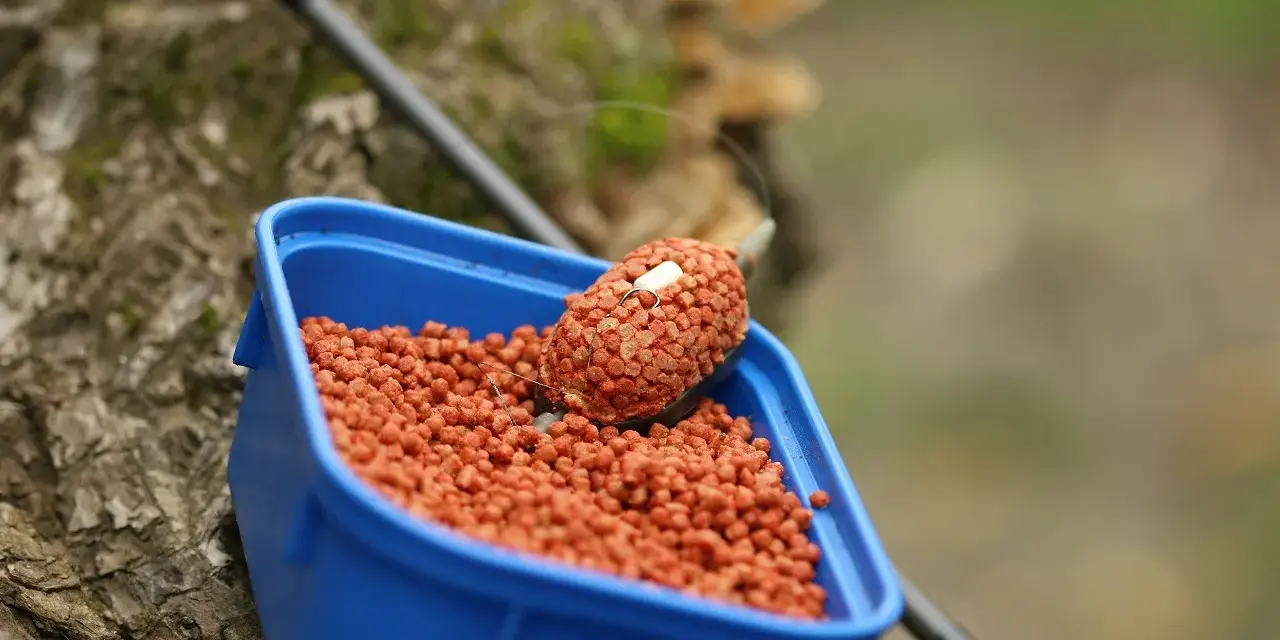 Zbliżenie na niebieskie pudełko z pomarańczowym pelletem, idealnym jako jaki pellet do metody na karpia. Na wierzchu znajduje się podajnik z przynętą.